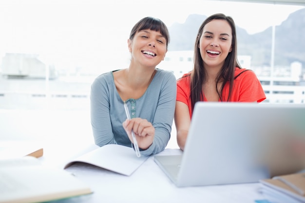 Due ragazze ridendo seduti davanti a un computer portatile insieme mentre guardano la telecamera