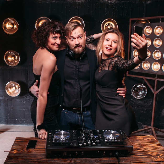 Due ragazze sexy adulte in abiti neri e un bel disc jockey barbuto che si fa selfie con lo smartphone alla console del DJ