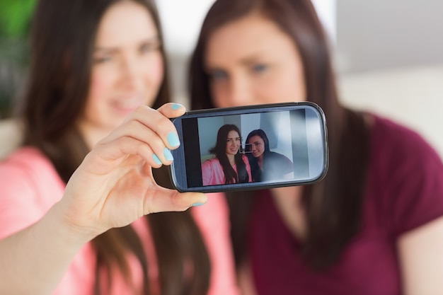 Due ragazze sorridenti che si siedono su un sofà che cattura una foto di se stessi con un telefono cellulare