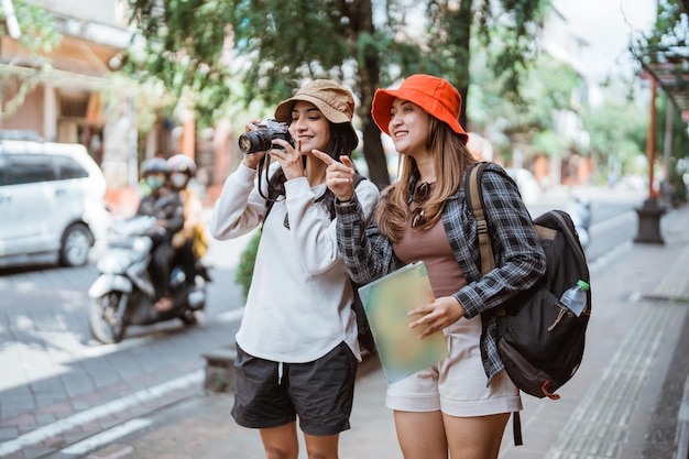 Due ragazze zaino in spalla camminano per trovare una destinazione turistica portando una fotocamera e una mappa sul ciglio della strada