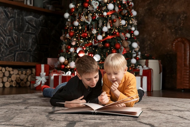 Due ragazzi leggono un libro sdraiato sul pavimento sotto l'albero di Natale I fratelli o gli amici scolaro leggono la storia di Natale o le fiabe