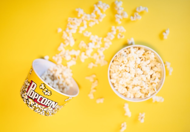 Due secchi pieni di popcorn uno ribaltato e circondato da molti popcorn su uno sfondo giallo