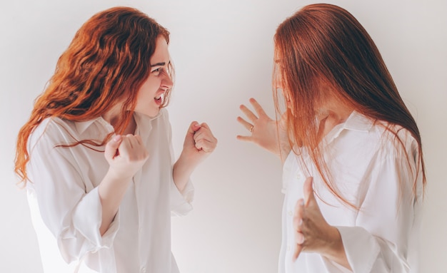 Due sorelle dai capelli rossi stanno isolate su uno sfondo bianco in ampie camicie di grandi dimensioni. Due giovani donne si arrabbiavano e si urlavano l'un l'altro Sentono rabbia, aggressività, rabbia