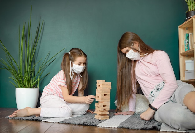 Due sorelle nella mascherina medica che gioca giocando un gioco di legno della torre dei blocchi