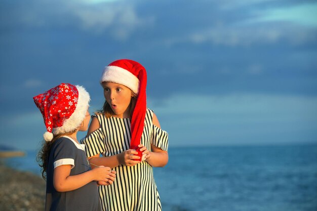 Due sorelle ragazze in cappelli di Babbo Natale si divertono in riva al mare al tramonto