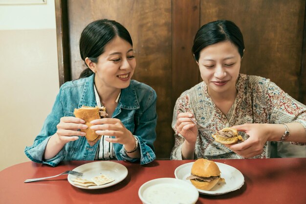 due sorelle sorridenti sedute al venditore locale in olvera street divertendosi durante il pranzo. gioia le giovani donne viaggiatori asiatici che provano cibo messicano nel ristorante del mercato all'aperto mangiano hamburger e panini