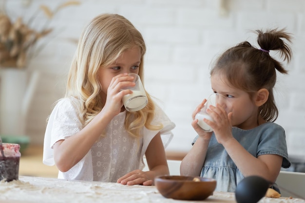 Due sorelline che bevono latte ecologico da bicchieri su un tavolo ricoperto di farina in cucina Felici fratelli di 5 e 8 anni che fanno una pausa e si godono del sano latte di mucca dopo aver fatto gustosi biscotti