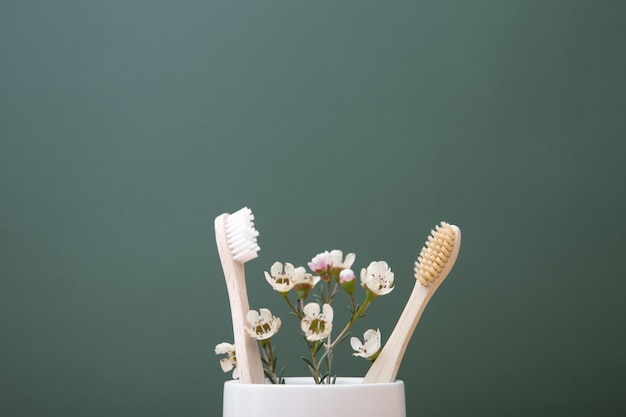 Due spazzolini da denti in legno di bambù e fiori in un bicchiere bianco su sfondo scuro Il concetto di sostenibilità e il rifiuto della plastica Il concetto di igiene e routine quotidiana Copia spazio
