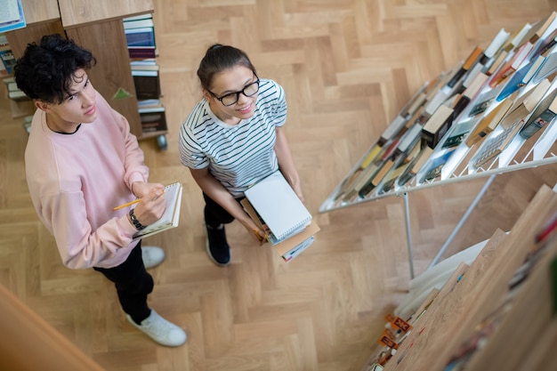 Due studenti adolescenti intelligenti in abbigliamento casual guardando uno degli scaffali nella biblioteca del college mentre si preparano per il seminario