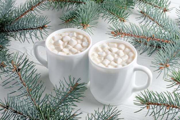 Due tazze bianche di cioccolata calda con marshmallow e albero di Natale su un tavolo di legno bianco. Bevanda al cacao in una mattina con decorazioni di Capodanno, rami di abete. Stagione invernale, card dal design minimale.