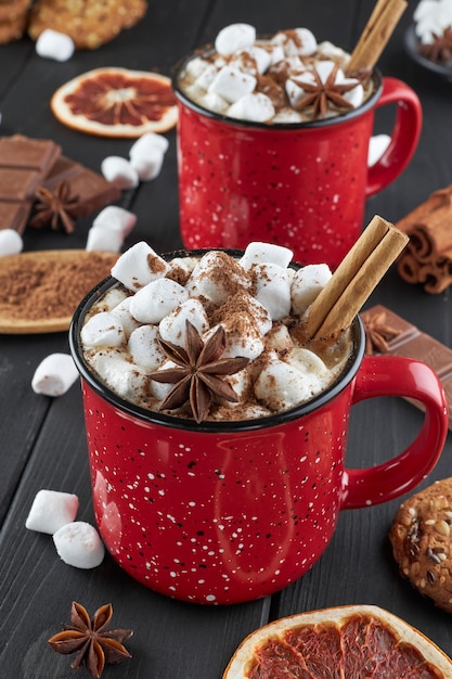 Due tazze rosse di cioccolata calda con marshmallow, anice e cannella cosparse di cacao in polvere