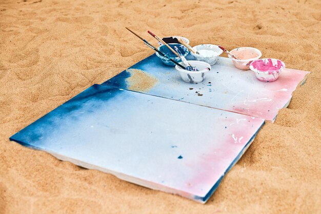 Due tele dipinte nei colori blu e rosa con tavolozze di colori sdraiato sulla spiaggia sabbiosa tavolozza artist39s al festival d'arte all'aperto Tela bianca da disegno con vernice in ciotole concetto di performance artistica