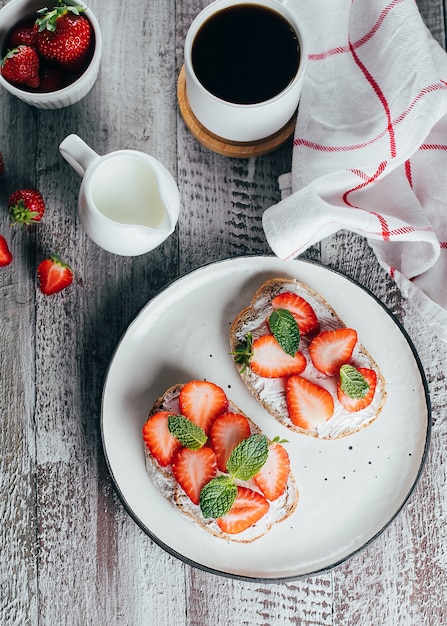 due toast o bruschette sul piatto con fragole su crema di formaggio e tazza di caffè sul tavolo di legno