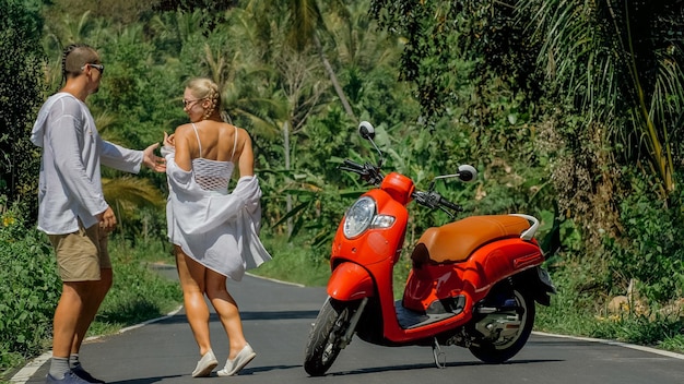 Due turisti caucasici donna uomo ballano vicino allo scooter. Coppia d'amore su una moto rossa in abiti bianchi, occhiali da sole durante il viaggio su strada forestale. Strada danzante. Noleggio moto. Asia Thailandia giro turistico.