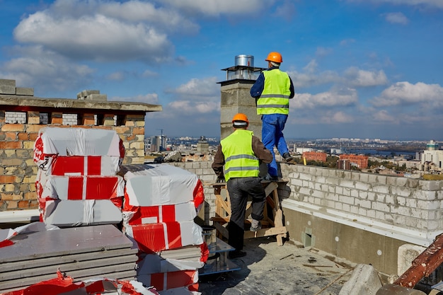 Due uomini costruttori professionisti in abiti da lavoro e elmetti protettivi arancioni stanno lavorando con un sistema di ventilazione sul tetto dell'edificio in costruzione