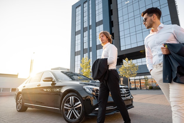 Due uomini d'affari camminano verso un'auto nera di lusso vicino all'hotel o all'edificio degli uffici al tramonto Concetto di trasporto e stile di vita aziendale