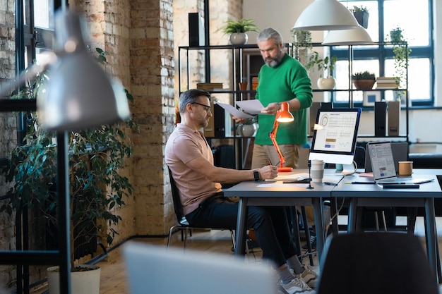 Due uomini d'affari che collaborano tra loro in un ufficio moderno utilizzando laptop e documenti che si preparano per la presentazione