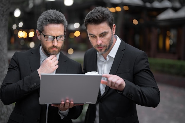 Due uomini d'affari che utilizzano il computer portatile uomini d'affari all'aperto all'aperto vicino al paesaggio urbano della città utilizzando il computer portatile