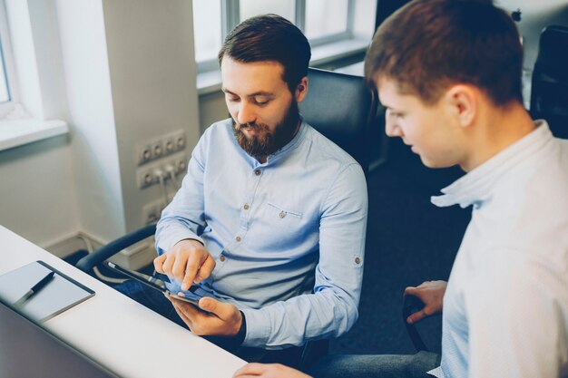 Due uomini in camicie eleganti seduti alla scrivania in ufficio e navigando insieme nel tablet moderno. Colleghi maschi che utilizzano tablet insieme