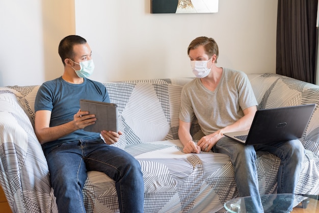 Due uomini multietnici con maschera come amici che usano la tecnologia e parlano in quarantena a casa