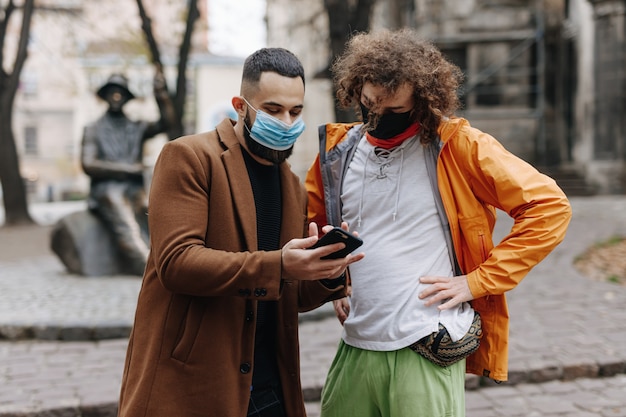 Due uomini multietnici in maschere mediche in piedi sulla strada e utilizzando smartphone. Concetto di persone, tecnologia e coronavirus.