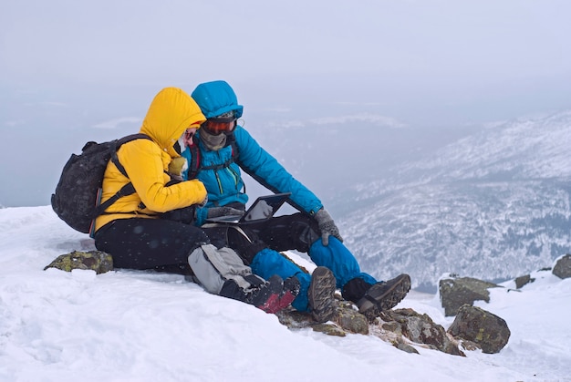 Due viaggiatori sono seduti con un laptop su una montagna invernale contro un paesaggio sfocato dell'altopiano