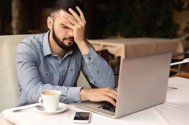 Duro lavoro a casa, mal di testa. L'uomo infelice tiene la testa mentre lavora su un computer portatile all'interno