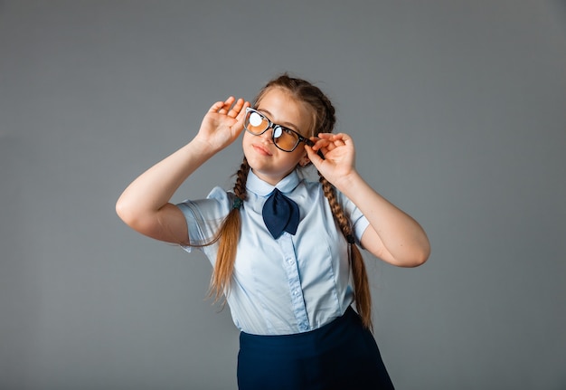 Eccitata bambina in uniforme scolastica e occhiali che si diverte e mostra dieci dita mentre sta in piedi su uno sfondo grigio