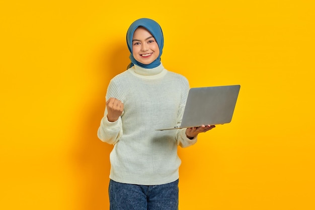 Eccitata bella donna asiatica in maglione bianco che tiene laptop e celebra la vittoria isolata su sfondo giallo persone concetto di stile di vita religioso