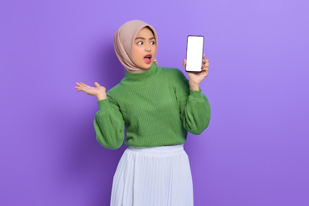 Eccitata bella donna asiatica in maglione verde e hijab che mostra il telefono cellulare con schermo vuoto isolato su sfondo viola