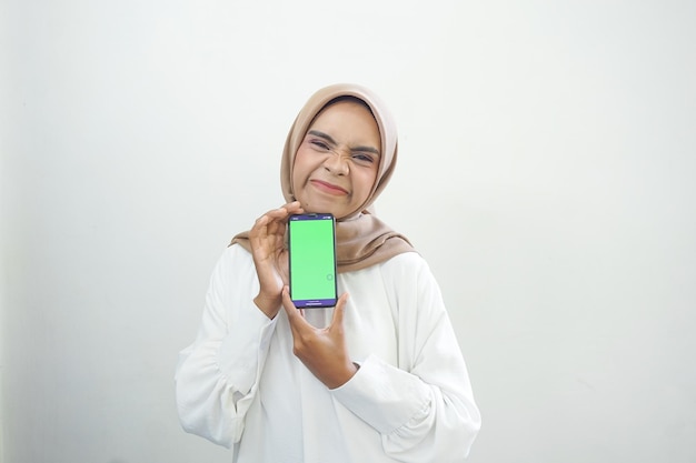 Eccitata bella donna musulmana asiatica che mostra il cellulare con schermo verde isolato su bianco