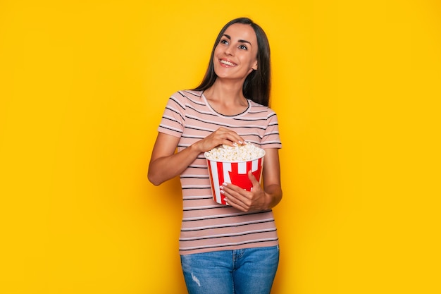 Eccitata bella e moderna giovane donna bruna in abbigliamento casual e occhiali da cinema 3d e grande cesto di popcorn è in posa mentre guarda un film al cinema