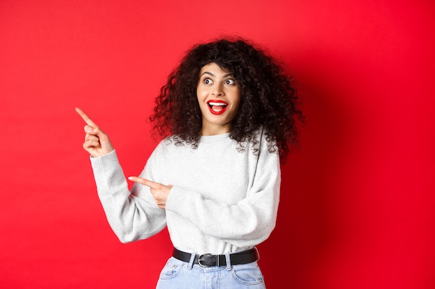 Eccitata bella ragazza con i capelli ricci e le labbra rosse, guardando e puntando le dita a sinistra con la faccia stupita, mostrando la bandiera, in piedi sul fondo dello studio.