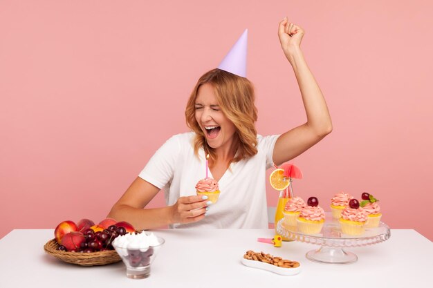 Eccitata bionda giovane donna adulta festeggia il compleanno tenendo la torta con la candela che urla felicemente godendosi il sogno che diventa realtà durante la sua vacanza Studio indoor girato isolato su sfondo rosa