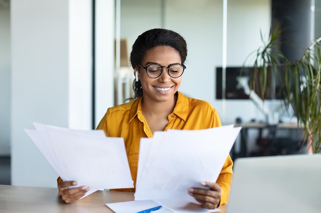 Eccitata donna d'affari nera che legge documenti e relazione d'affari positiva seduta sul posto di lavoro nell'interiore dell'ufficio moderno