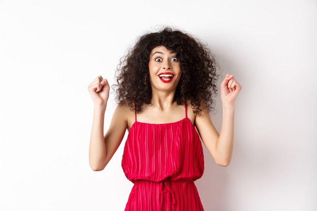 Eccitata donna dai capelli ricci con trucco da sera, ansimante affascinata, alzando le mani e gioendo, vincendo un premio e festeggiando, in piedi su sfondo bianco.