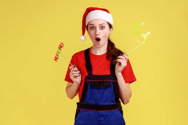 Eccitata donna di consegna felice con oggetti di scena per feste di Natale che urlano di eccitazione, festeggia il nuovo anno, indossando tute blu e cappello di Babbo Natale. Studio indoor girato isolato su sfondo giallo.