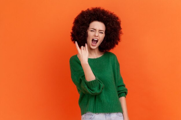 Eccitata donna entusiasta con acconciatura afro che indossa un maglione verde in stile casual che mostra un gesto rock and roll che urla estaticamente in studio indoor girato isolato su sfondo arancione