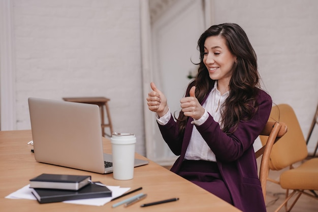 Eccitata donna ispanica castana in abito viola seduto alla scrivania con computer portatile e tazza di caffè