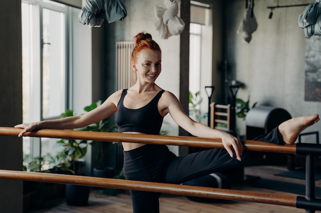 Eccitata donna magra dai capelli rossi in piedi in un moderno studio fitness con le mani e una gamba sulla sbarra da balletto, facendo esercizi di stretching e guardando nella telecamera, indossando un comodo abbigliamento sportivo nero