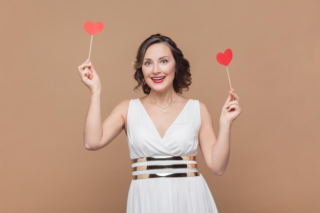 Eccitata donna positiva con capelli ondulati che tiene piccoli cuori rossi che sorridono felicemente festeggiando