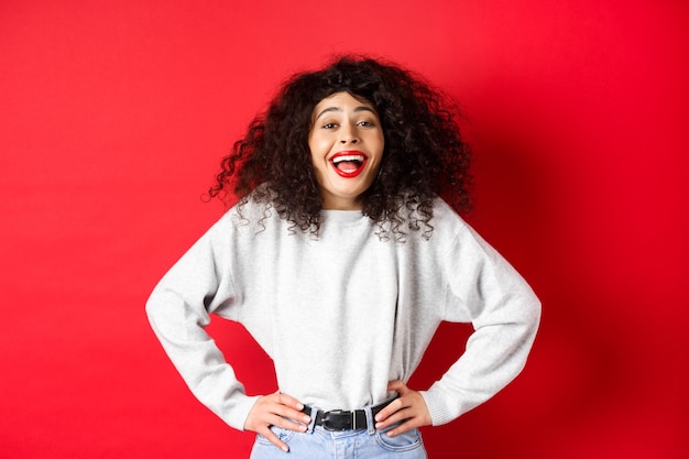 Eccitata donna positiva ridendo e sorridendo, sentendosi ottimista, guardando il promo divertente, in piedi su sfondo rosso.