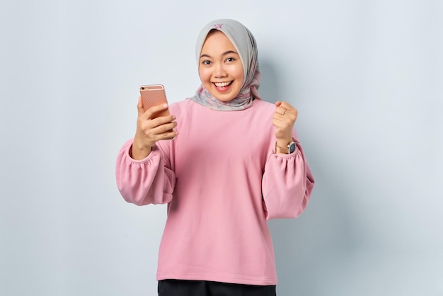 Eccitata giovane donna asiatica in camicia rosa utilizzando il telefono cellulare ha ricevuto buone notizie isolate su sfondo bianco