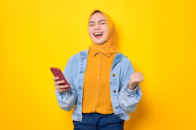 Eccitata giovane donna asiatica in giacca di jeans utilizzando il telefono cellulare per celebrare il successo ottenendo buone notizie isolate su sfondo giallo