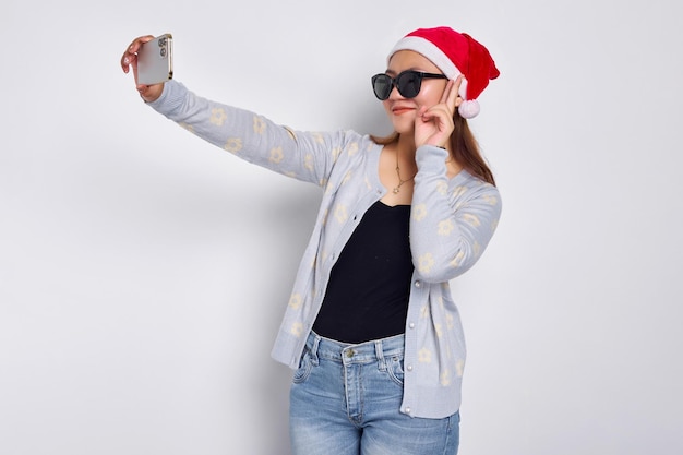 Eccitata giovane donna asiatica in un cappello di Natale che fa un selfie girato su un telefono cellulare mobile pubblica una foto sul social network isolato su sfondo bianco