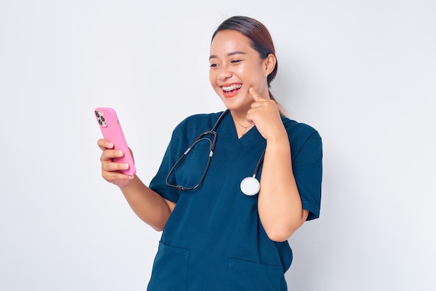 Eccitata giovane donna asiatica infermiera che lavora indossando un'uniforme blu guardando lo schermo dello smartphone e reagendo emotivamente alle notizie online isolate su sfondo bianco Concetto di medicina sanitaria