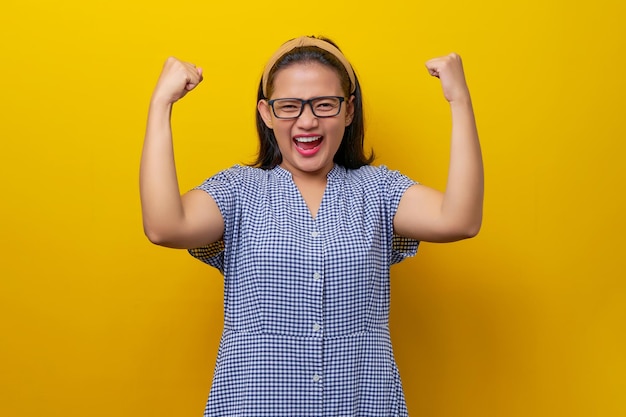 Eccitata giovane donna asiatica soddisfatta che indossa un abito a scacchi con gli occhiali alzando i pugni celebrando il successo isolato su sfondo giallo concetto di stile di vita delle persone