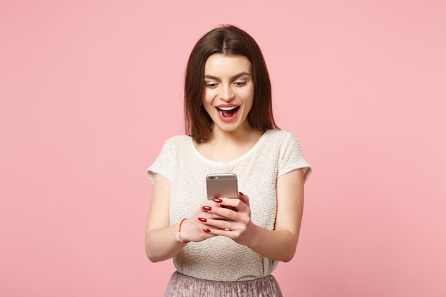 Eccitata giovane donna gioiosa in abiti leggeri casual in posa isolata su sfondo rosa pastello parete ritratto in studio. Concetto di stile di vita delle persone. Mock up spazio di copia. Utilizzando il telefono cellulare digitando un messaggio sms.