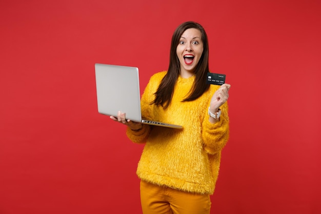 Eccitata giovane donna in maglione di pelliccia gialla che tiene la bocca spalancata, tiene in mano una carta di credito, un computer portatile isolato su sfondo rosso. Persone sincere emozioni, concetto di stile di vita. Mock up copia spazio.