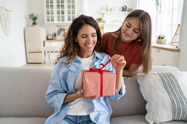 Eccitata giovane donna madre che si fa avvolgere confezione regalo presente dalla figlia mentre si siede sul divano